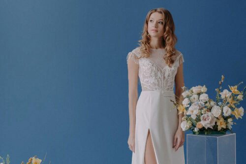 Braut in einem eleganten Kleid steht vor einem blauen Hintergrund neben einem Blumenarrangement.