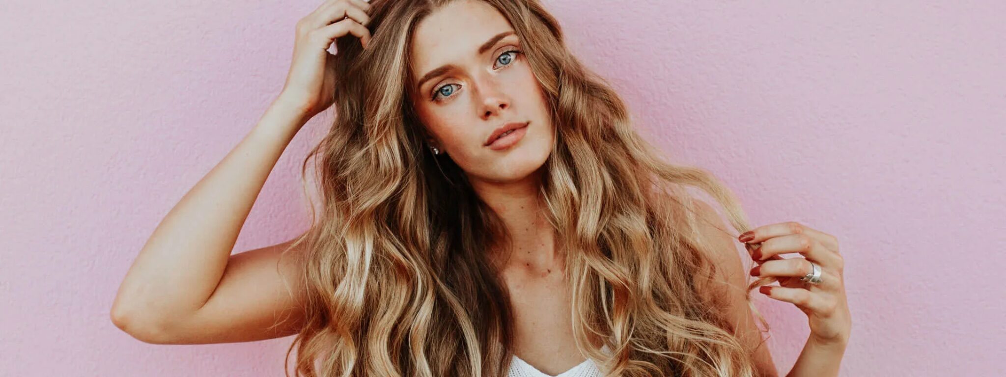 Junge Frau mit langen, voluminösen Locken vor einem rosafarbenen Hintergrund, hält eine Hand in ihr Haar.