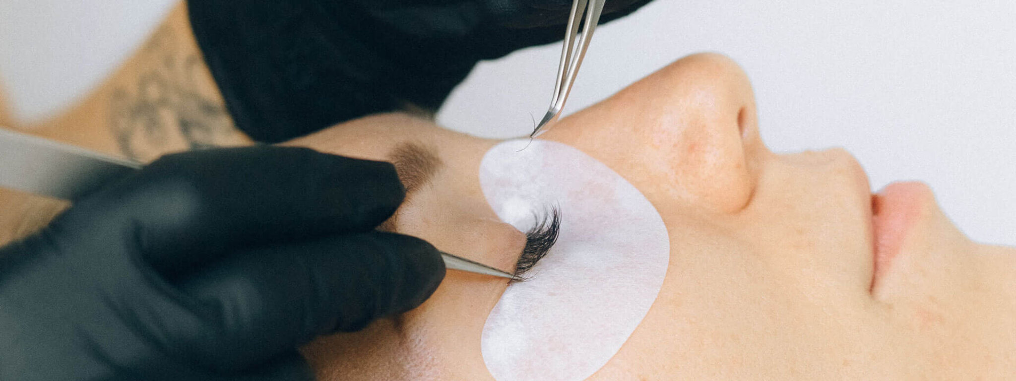 Frau während einer Wimpernverlängerung, Kosmetikerin in schwarzen Handschuhen trägt mit einer Pinzette einzelne Wimpern auf, Augen mit Schutzpads abgedeckt.