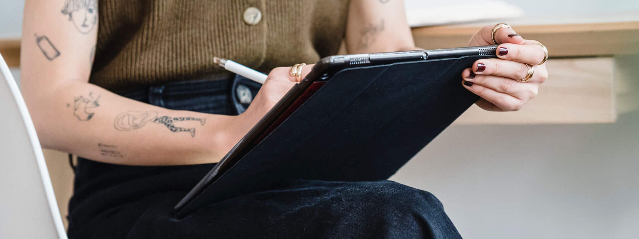 Person in ärmellosem Oberteil und dunkler Hose, sitzend auf einem Stuhl und schreibend auf einem Klemmbrett, mit sichtbaren Tattoos auf den Armen