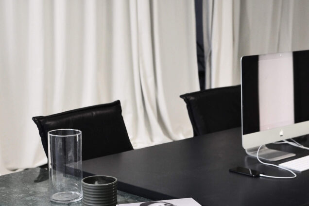Moderner Arbeitsplatz mit einem Computer, Bücherstapel, Vasen und einer Tasse auf einem Marmortisch, vor einem beigen Vorhang.
