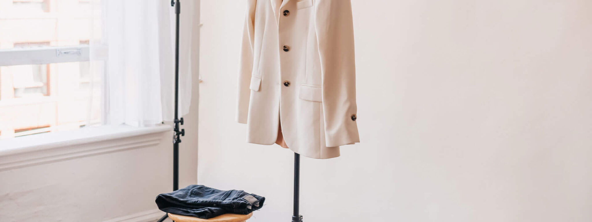 Fotostudio-Setup mit beigefarbenem Hintergrund, einem hellen Blazer an einem Ständer, Jeans auf einem Hocker und Accessoires auf einem kleinen Tisch.