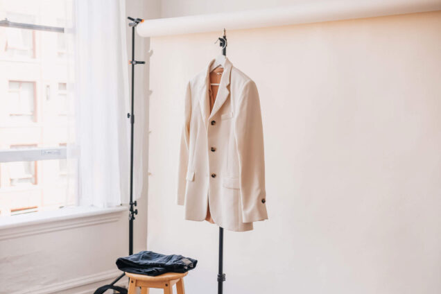 Fotostudio-Setup mit beigefarbenem Hintergrund, einem hellen Blazer an einem Ständer, Jeans auf einem Hocker und Accessoires auf einem kleinen Tisch.
