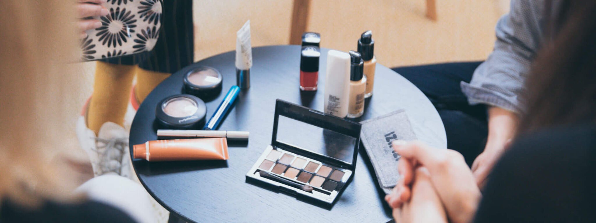 Gruppe von Menschen, die sich um einen Tisch mit verschiedenen Make-up-Produkten versammelt haben, darunter Lidschatten, Lippenstifte und Pinsel.
