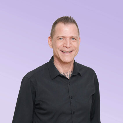 Porträt eines lächelnden Mannes in einem schwarzen Hemd vor einem hellvioletten Hintergrund.