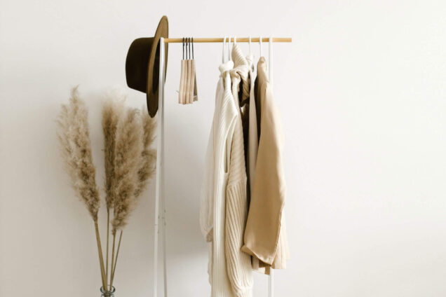 Minimalistische Kleiderstange mit beige- und cremefarbenen Kleidungsstücken, daneben eine große Vase mit Pampasgras und eine geflochtene Tasche auf dem Boden