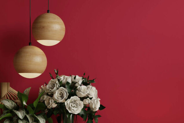 Blumenstrauß in einer Vase auf einem Holztisch vor einer dunkelroten Wand, dekoriert mit zwei hängenden, runden Holzlampen
