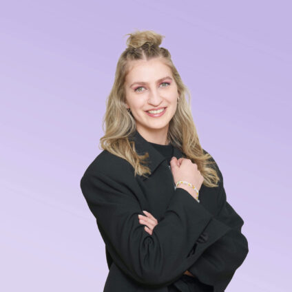 Porträt einer lächelnden Frau mit langen blonden Haaren, die teilweise hochgesteckt sind, in einem schwarzen Blazer vor einem hellvioletten Hintergrund