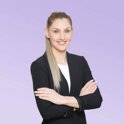 Porträt einer lächelnden Frau mit langen, glatten blonden Haaren, die die Arme verschränkt hat, in einem schwarzen Blazer vor einem hellvioletten Hintergrund.