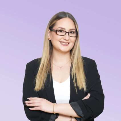 Porträt einer lächelnden Frau mit langen blonden Haaren und Brille, in einem schwarzen Blazer vor einem hellvioletten Hintergrund.