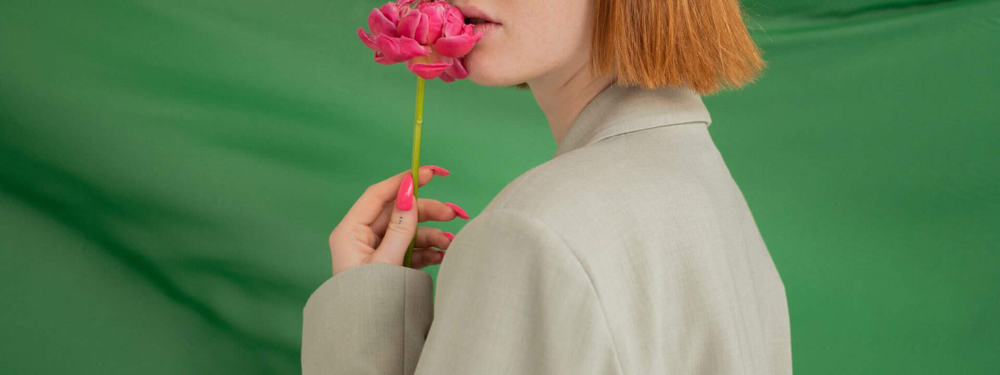 Frau mit kurzem, rötlichem Haar hält eine rosa Blume vor sich, während sie vor einem grünen Hintergrund steht.