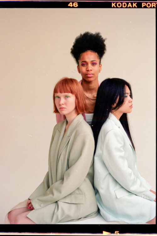 Drei Frauen in pastellfarbenen Outfits sitzen nebeneinander, vor einem hellen Hintergrund.